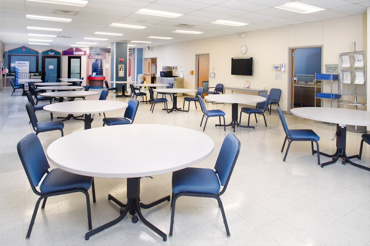 An empty cafeteria with round tables and blue chairs, well-lit by ceiling lights.