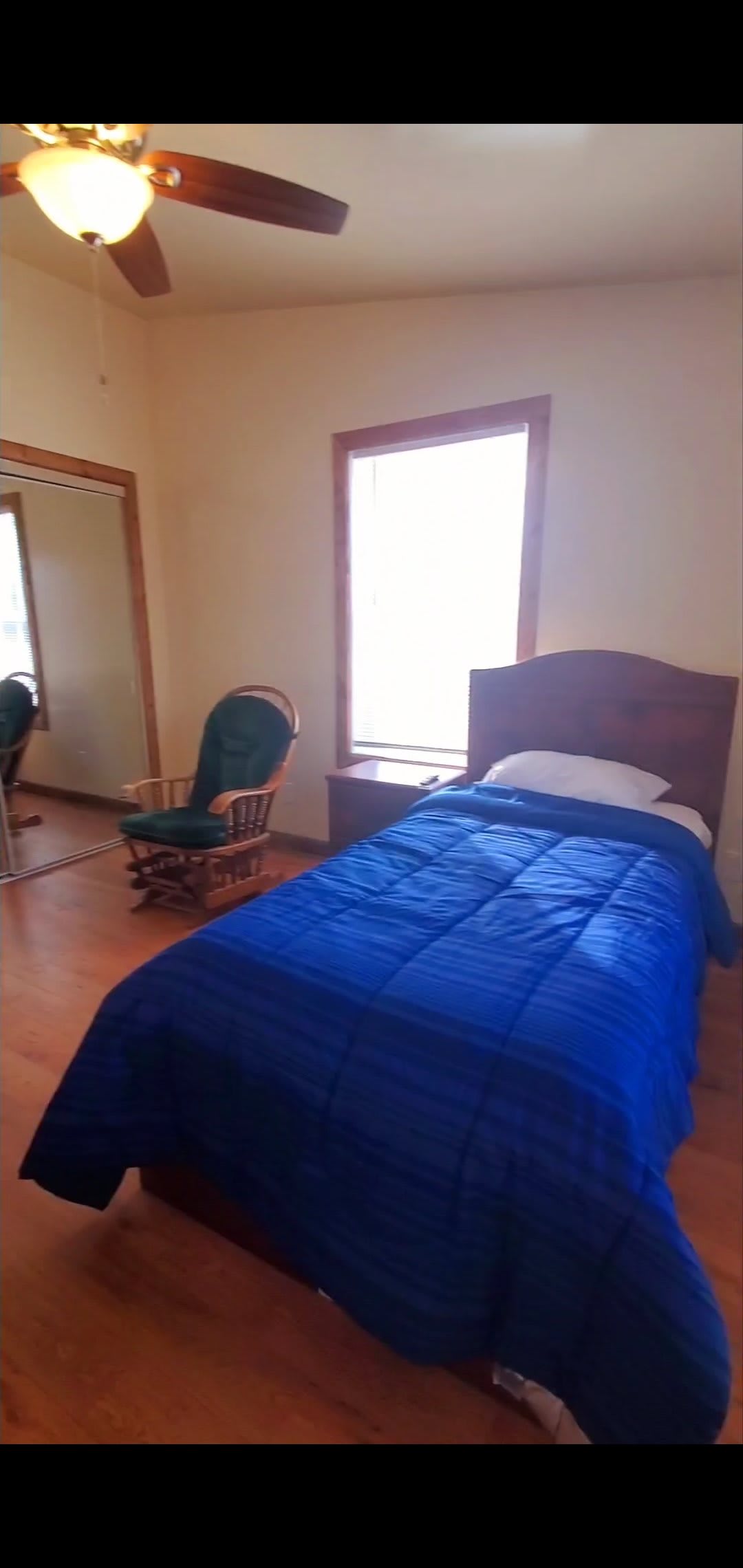 A minimalist bedroom featuring a single bed with a blue blanket, a rocking chair, and wooden flooring.