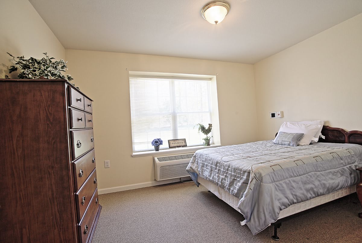 A simple bedroom with a bed, dresser, and window, featuring neutral colors and minimalist decor.