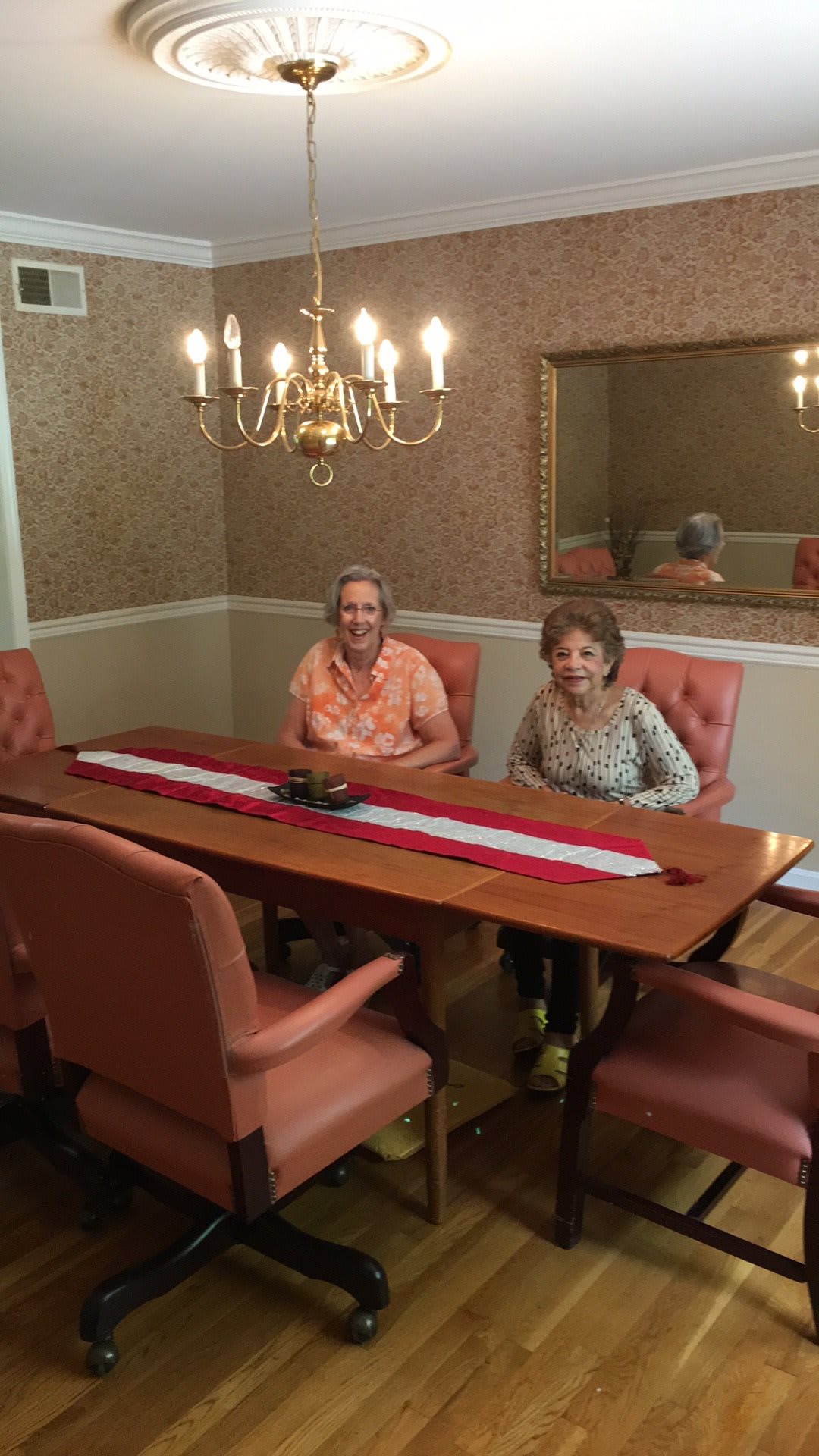 Two individuals seated at a dining table with a chandelier above and a mirror on the wall.