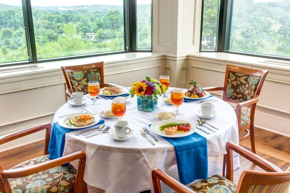 A beautifully set dining table with a floral centerpiece and food, overlooking a scenic view through large windows.