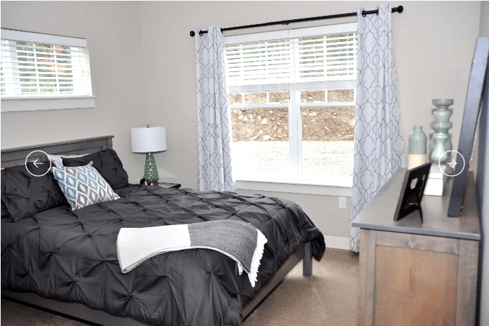 A modern bedroom with a neatly made bed, featuring dark bedding and patterned pillows. A lamp sits on the side table next to the window with light curtains.