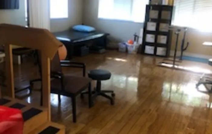 A cozy room with wooden flooring, featuring a bed, chairs, shelves, and ample natural light.