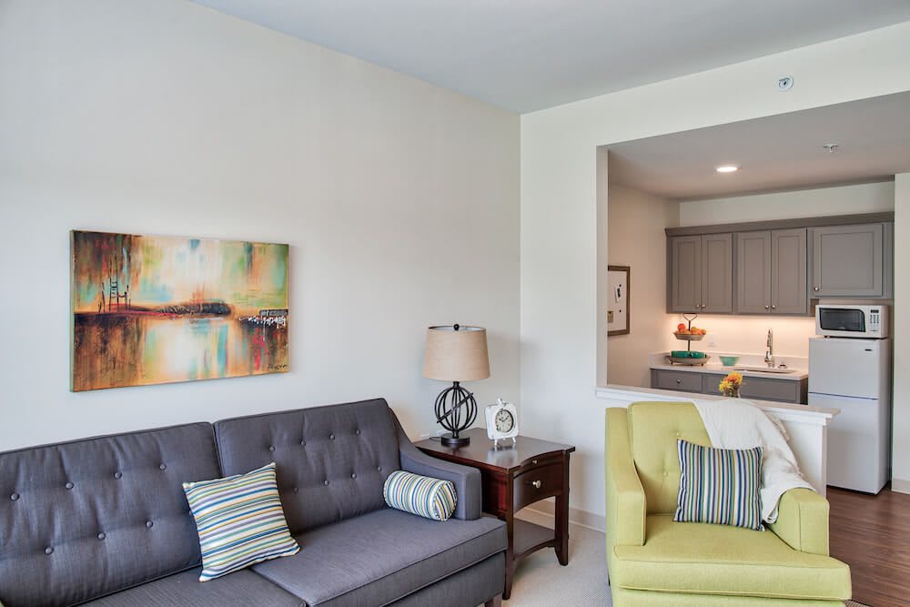 A cozy living room with a gray sofa and yellow armchair, featuring modern artwork and a view into the kitchen.