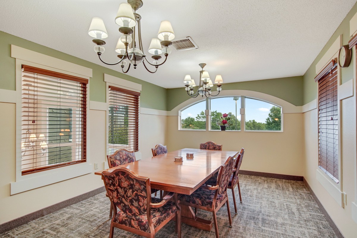 A well-lit dining room with a wooden table and six chairs, featuring chandeliers and large windows.