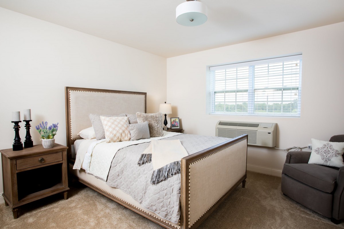 A cozy and well-decorated bedroom featuring a large bed with plush pillows, a side table with a lamp, and a window allowing natural light in.