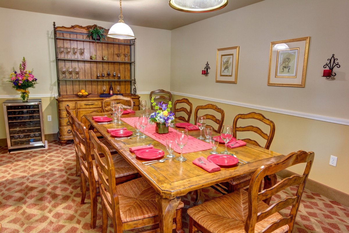 A warmly lit dining room with a large wooden table set for eight, featuring red placemats and a floral centerpiece.