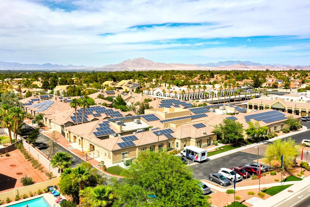 Aerial view of a residential area with solar panels on rooftops and a nearby parking lot, surrounded by trees and mountains in the background.
