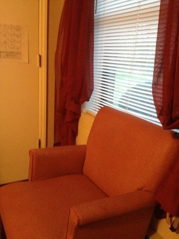 A cozy room corner featuring an orange armchair next to a window with blinds and curtains.