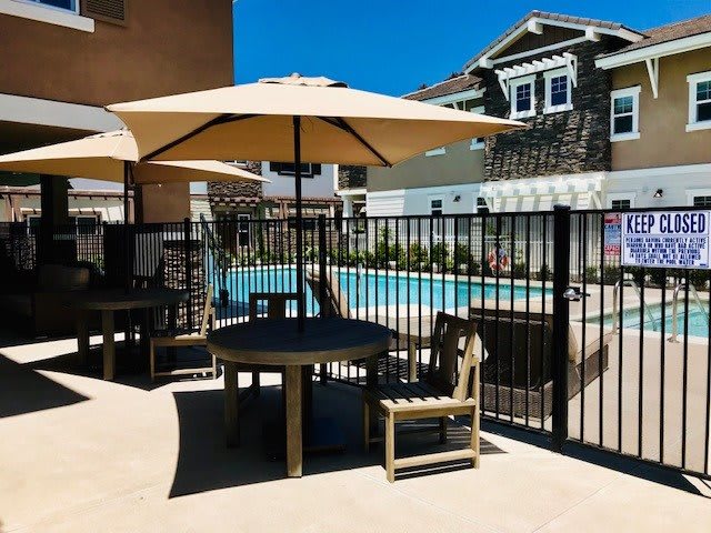 A sunny outdoor patio area with an umbrella-shaded table and chairs near a fenced swimming pool.