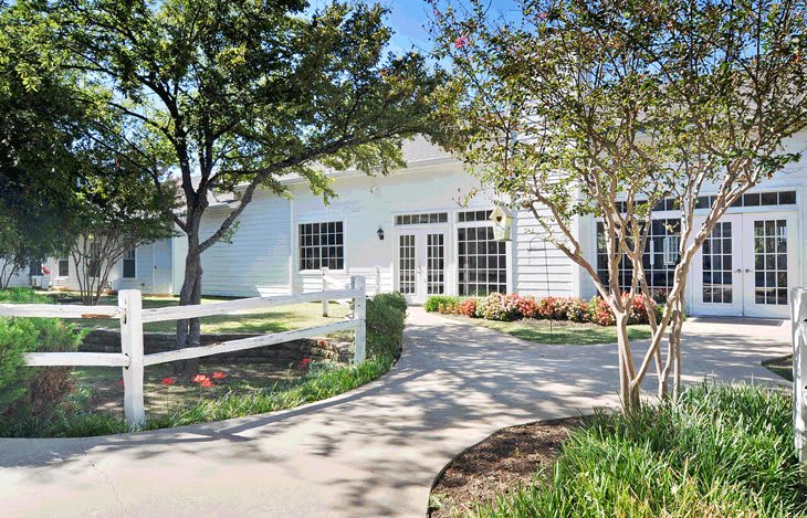 A charming white house with a winding pathway, surrounded by trees and well-kept shrubbery on a sunny day.