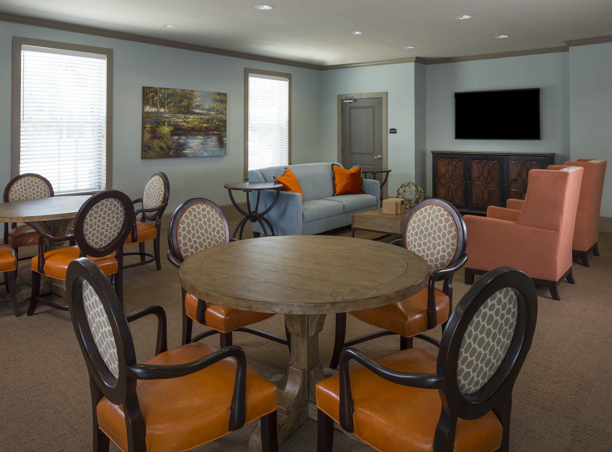 A cozy living room with a central table surrounded by chairs, a sofa with orange cushions, and a wall-mounted television.