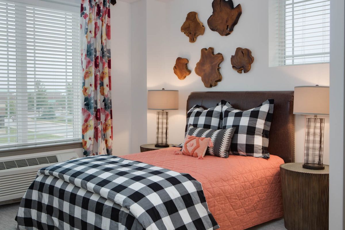 A cozy bedroom featuring plaid black and white bedding with decorative pillows, bright curtains, wooden wall art, and a warm table lamp.