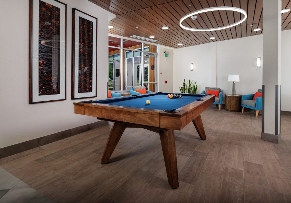 A modern game room featuring a pool table with wooden legs, surrounded by colorful seating and stylish pendant lighting.