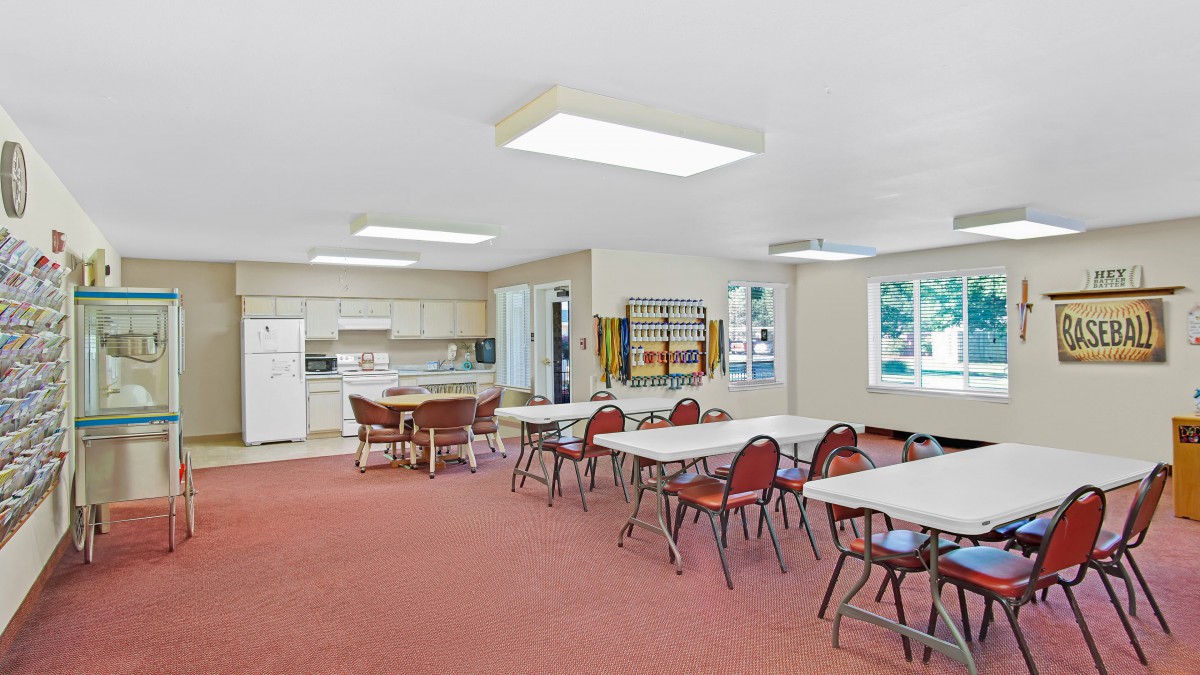 A community room featuring a kitchen area with tables and chairs, set on a carpeted floor with windows providing natural light.