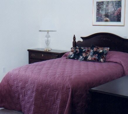 A neatly arranged bedroom with a purple quilt and floral pillows.