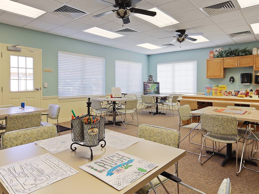 A well-lit classroom with multiple tables and chairs, ceiling fans, and large windows.