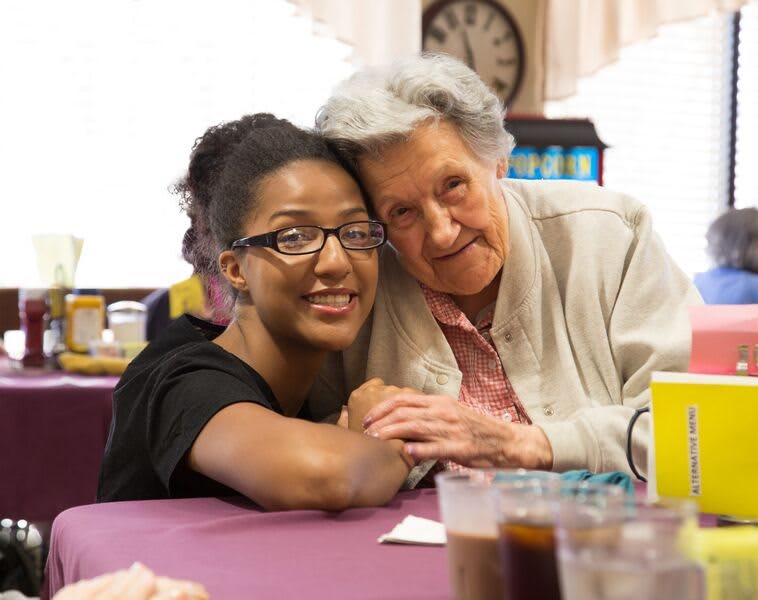 An elderly woman and a caregiver share a warm embrace, smiling at the camera.
