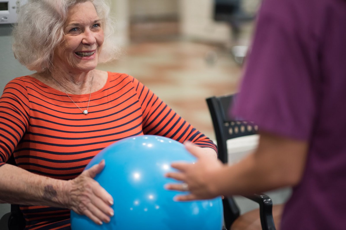 An elderly woman exercises with a therapy ball under guidance.