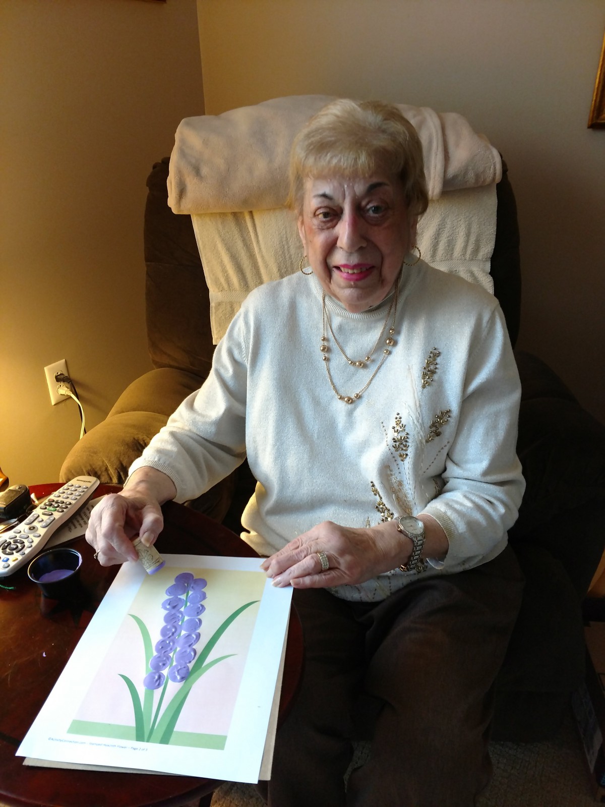 An elderly woman sitting indoors, holding a piece of floral artwork.