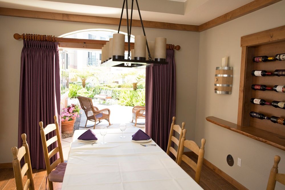 A cozy dining room with a white tablecloth, wooden chairs, and a view of the patio through purple curtains.