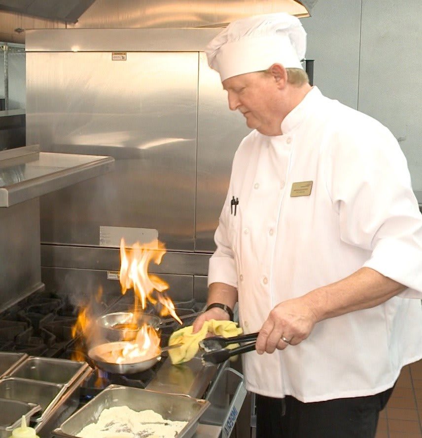 A chef expertly handles a dish on the stove with flames rising in a professional kitchen.