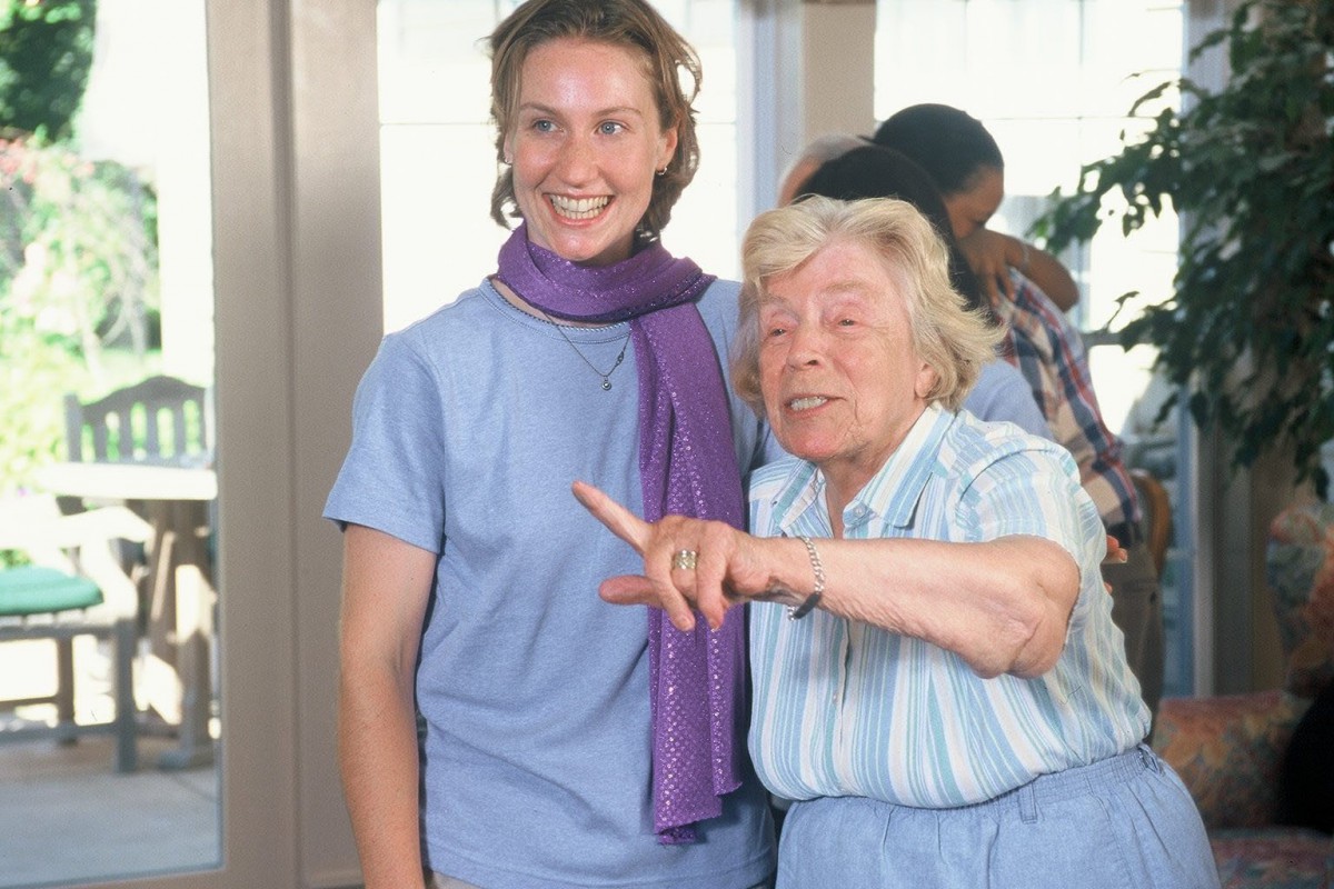 Two people are smiling and interacting indoors, dressed in casual clothing.