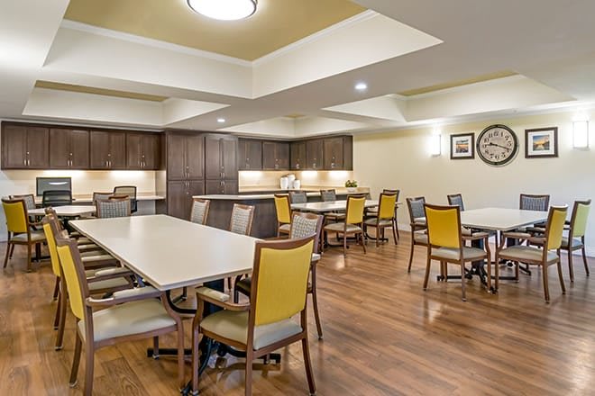 A spacious dining area with multiple tables and chairs, featuring wooden flooring and a kitchen section with cabinets.