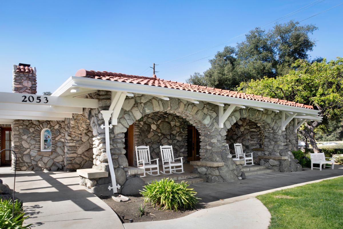 A charming stone house with a cozy porch featuring rocking chairs, surrounded by greenery.