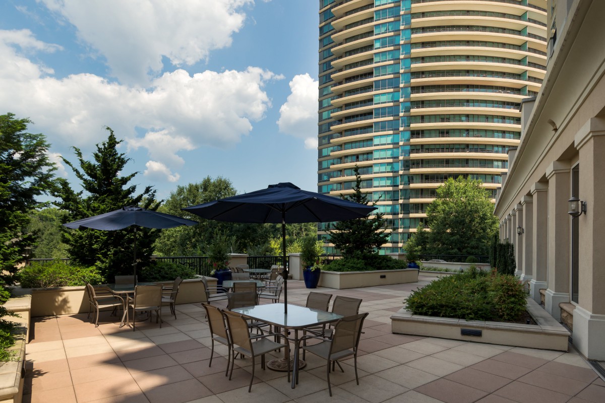 A modern patio with tables and umbrellas next to a high-rise building under a partly cloudy sky.