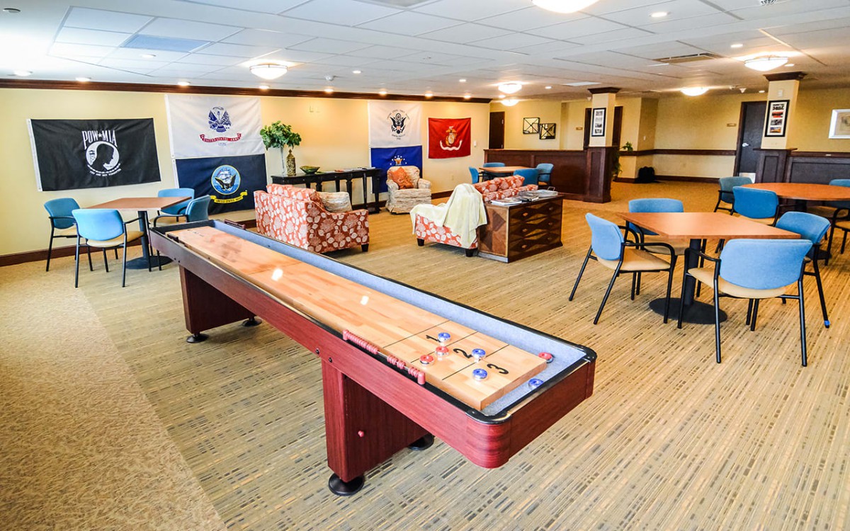 A spacious game room featuring a shuffleboard table, comfortable seating, and various flags on the wall.