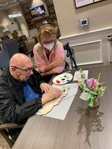 A caregiver assists an elderly man in painting, highlighting the importance of creative activities for mental well-being.