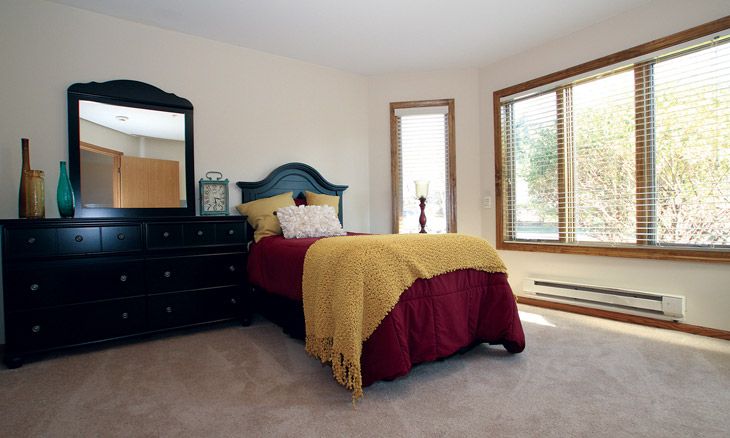 A cozy bedroom with a single bed adorned with burgundy and yellow bedding, featuring a dresser and large windows letting in natural light.
