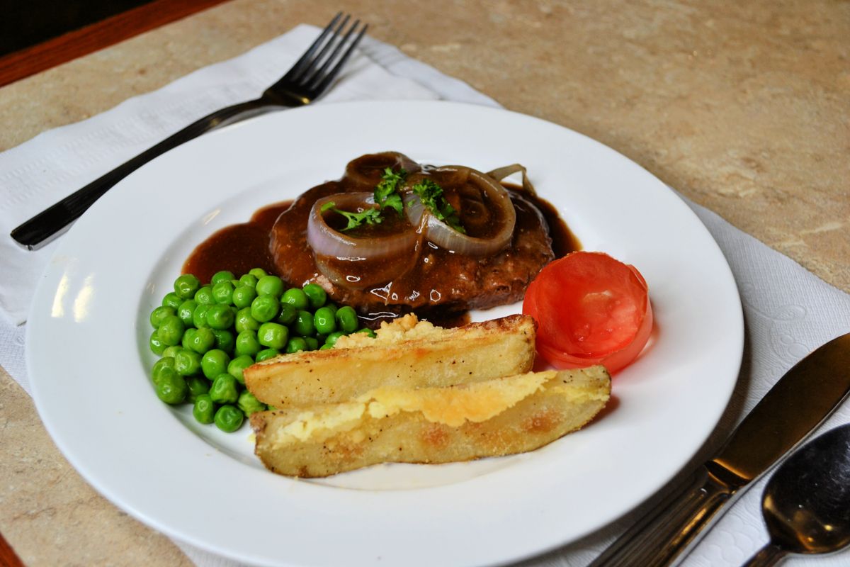 A plated meal featuring steak with gravy and onions, accompanied by peas, tomato, and potato wedges.