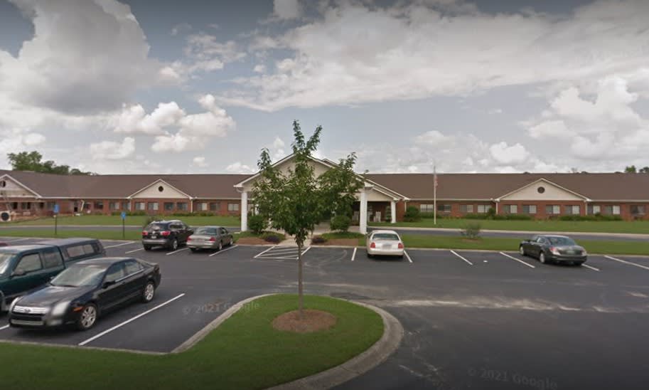 A single-story building with a parking lot in front, featuring several cars and a tree, under a cloudy sky.