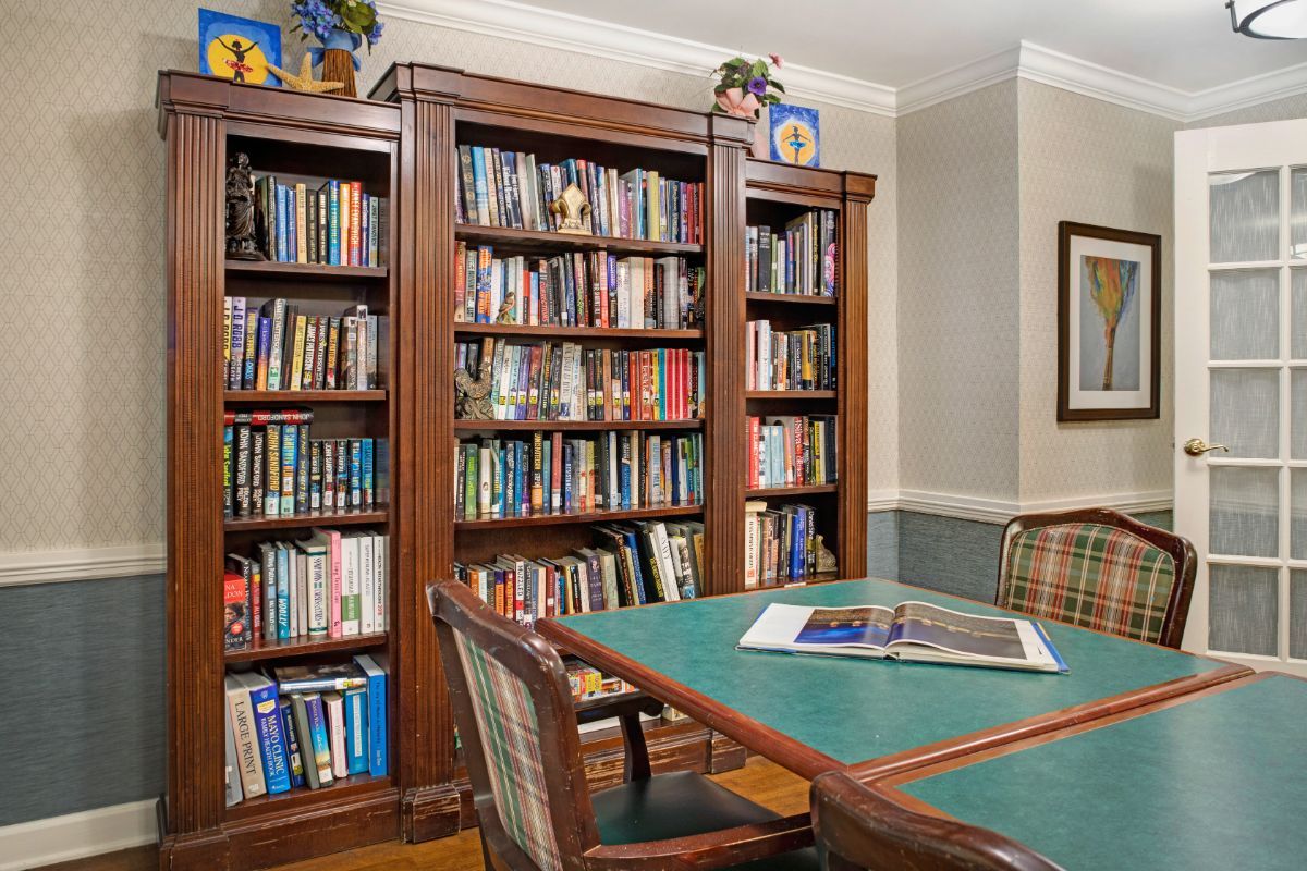 A cozy library room with wooden bookshelves filled with books and a central table surrounded by chairs.