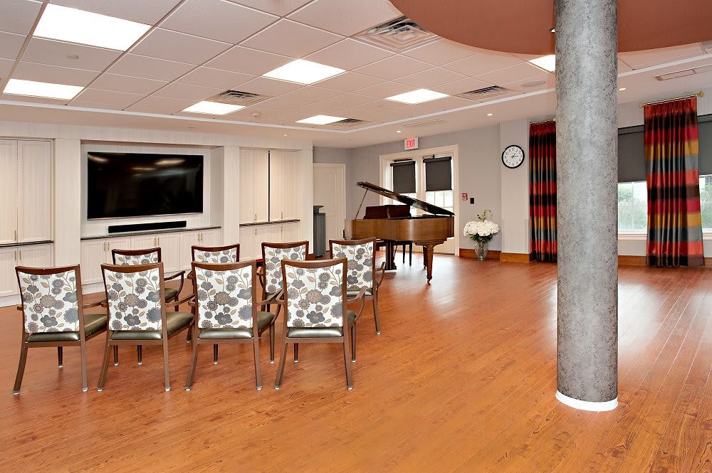 A modern room with chairs arranged facing a grand piano and a large television on the wall.