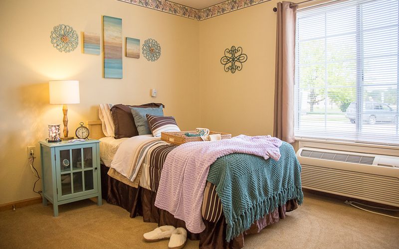 A cozy bedroom with pastel decor and soft bedding, featuring wall art and a large window.