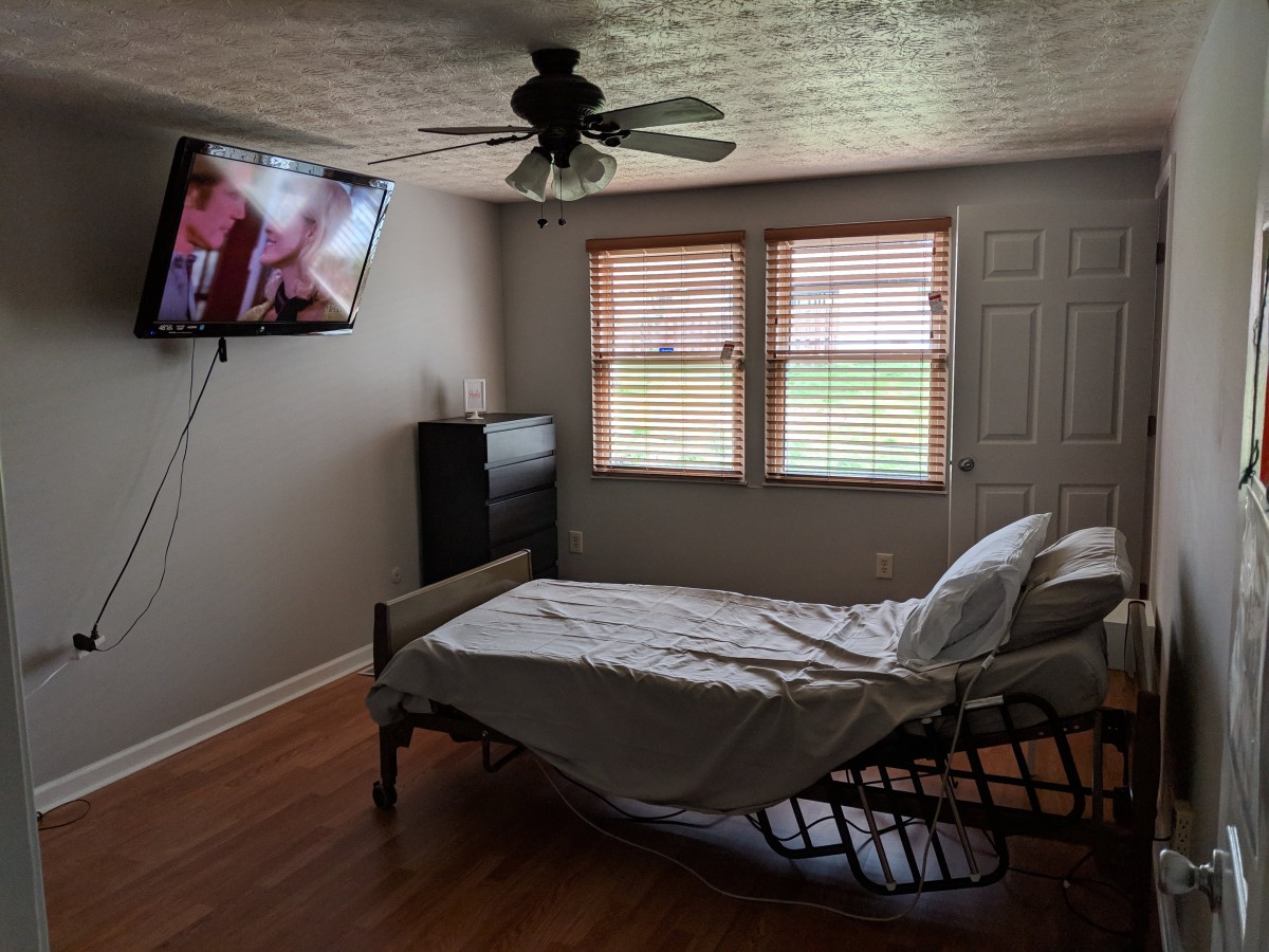 A bedroom with a hospital bed, wall-mounted TV, and windows with blinds.