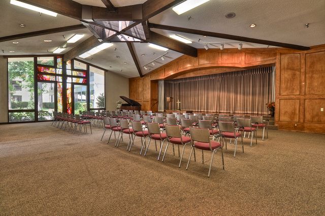 An empty auditorium with rows of chairs facing a stage and a piano near the window.