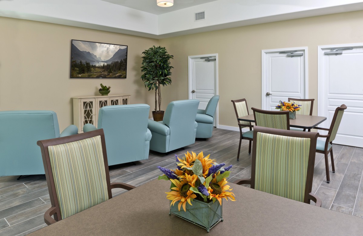 A cozy living room area with light blue armchairs, green striped chairs, tables decorated with flower arrangements, and a landscape painting on the wall.
