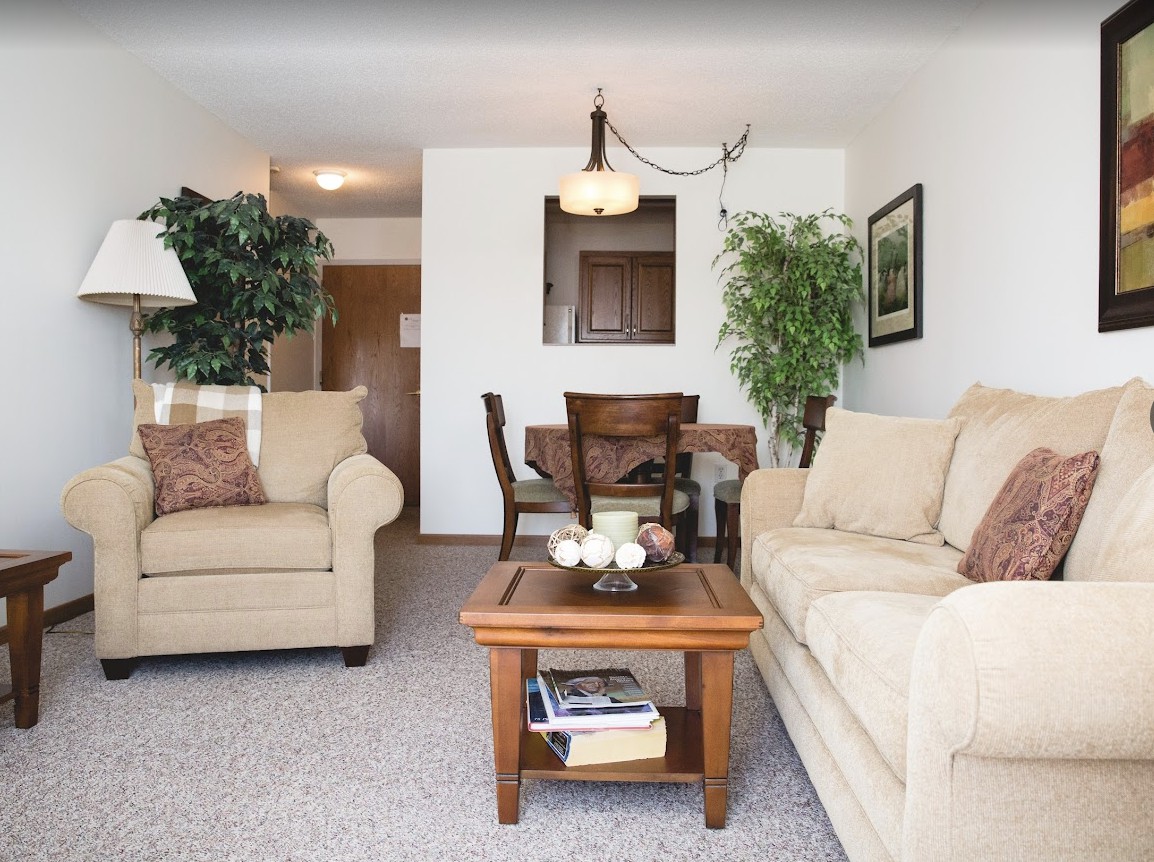 A cozy living room with beige sofas, a wooden coffee table, and green plants, adjacent to a dining area.