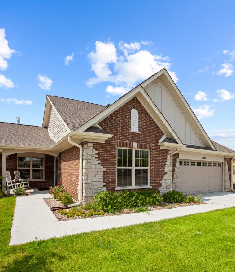 A suburban brick house with a garage and well-maintained lawn under a blue sky with clouds.