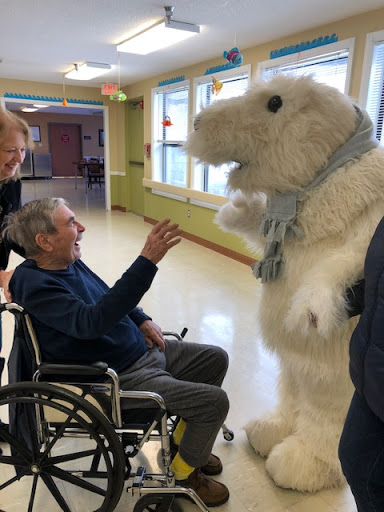 A joyful interaction between an elderly man in a wheelchair and a person dressed in a fluffy animal costume, bringing smiles to the healthcare setting.