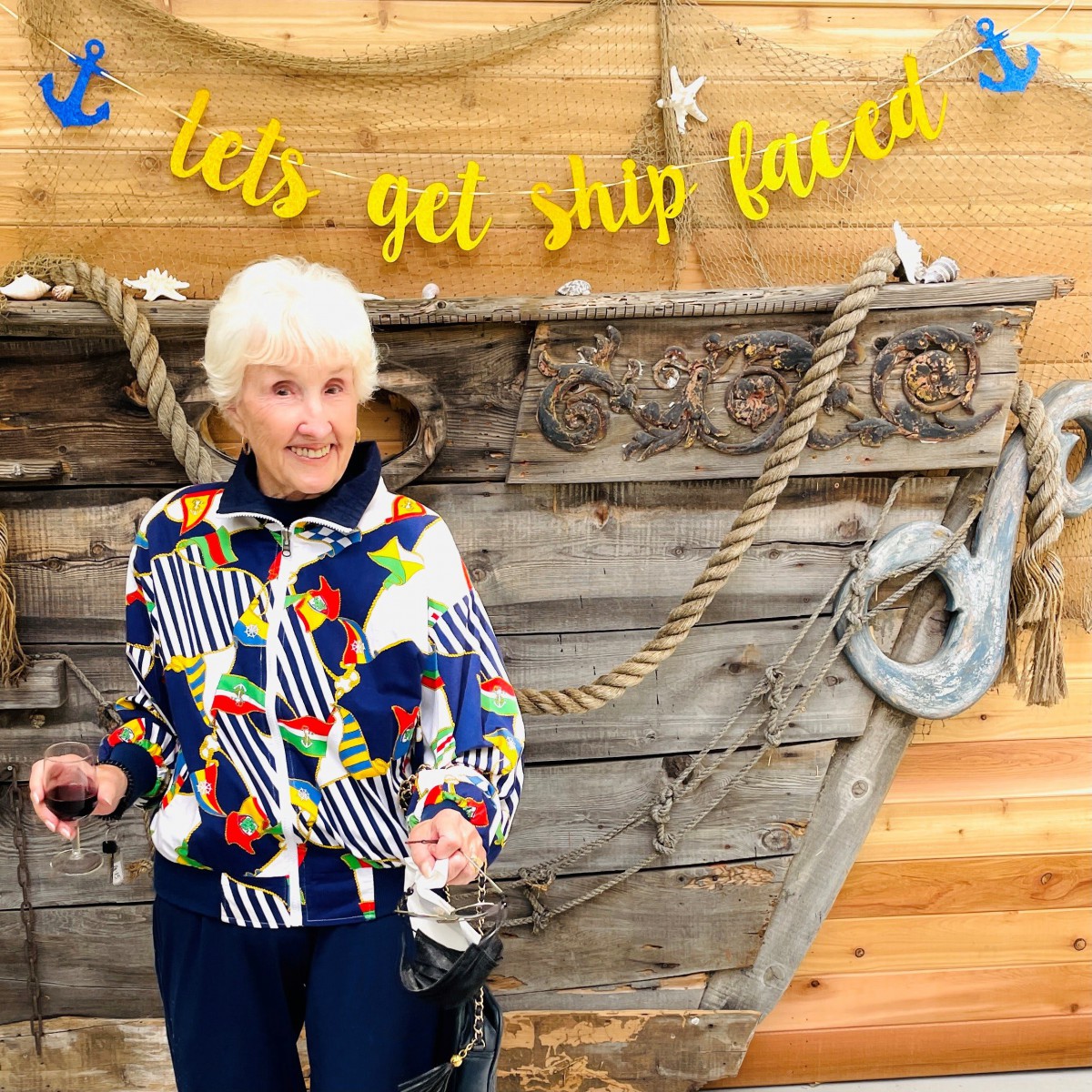An elderly woman stands in front of a decorative ship with a colorful jacket, under a banner that reads 'let's get ship faced'.