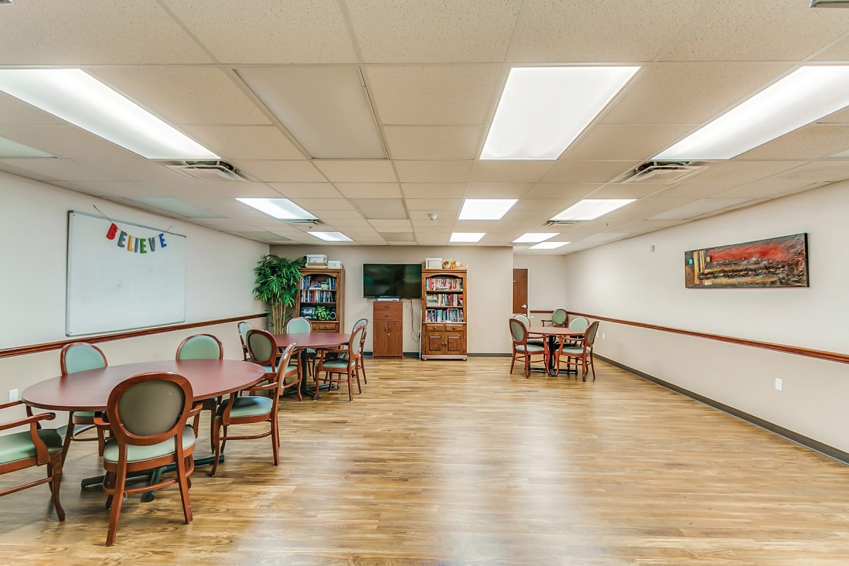 A spacious room with tables, chairs, and a television, featuring wooden flooring and bright lighting.