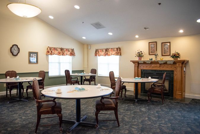 A cozy dining area with tables and chairs, featuring a fireplace and windows with decorative curtains.