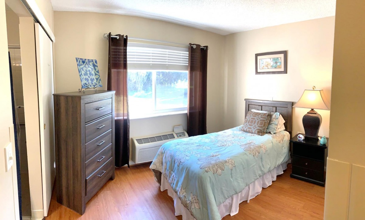 A cozy bedroom featuring a single bed with floral bedding, positioned beside a window adorned with dark curtains. A wooden dresser stands in the corner, completing the room's warm and inviting atmosphere.