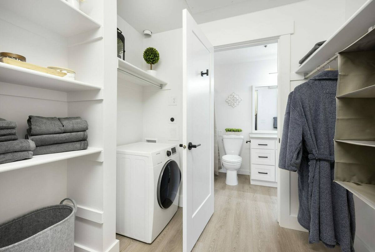 A modern laundry area connected to a bathroom, featuring white walls and shelves with neatly arranged towels.