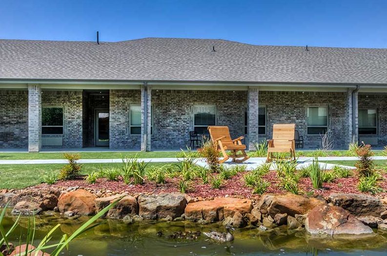 A backyard view featuring a house with patio chairs, a well-maintained lawn, and a picturesque pond with surrounding rocks.